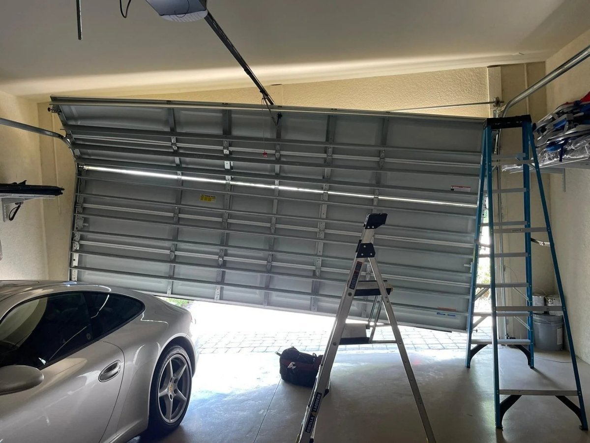 Sectional garage door sitting crooked above a parked car, with two ladders in the bay
